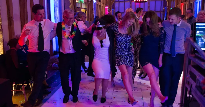 Wedding guests dancing arm in arm across a lit-up dance floor in a timber-framed barn
