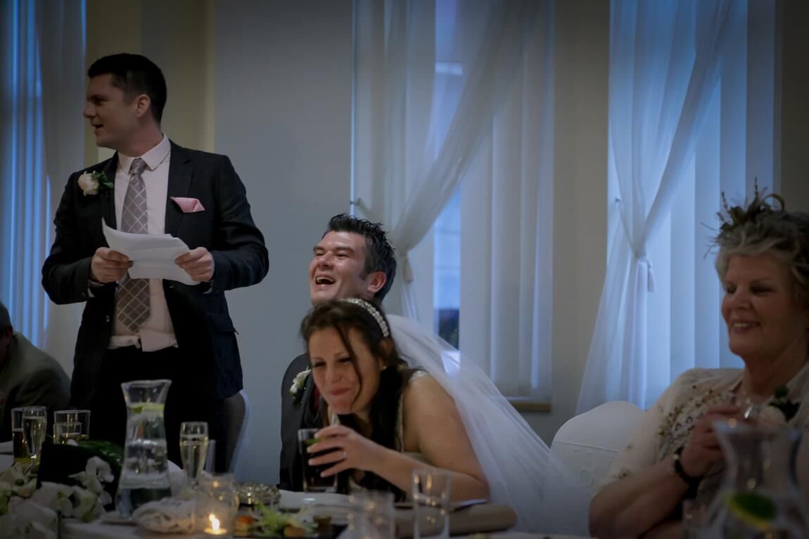 A bride and groom laughing at the top table as a guest reads his speech
