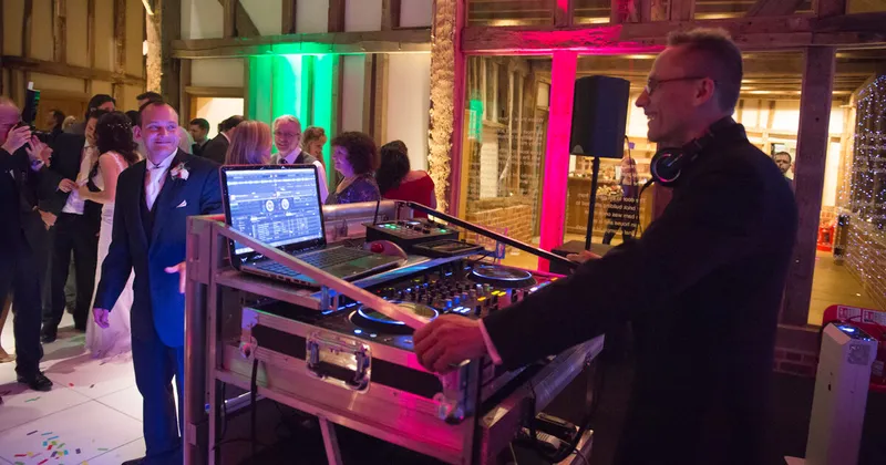 Tony smiling at the groom from behind his DJ booth as guests dance in a barn lit pink and green