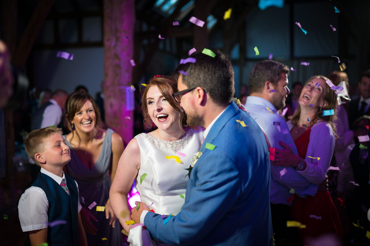 A bride and groom laughing as confetti falls during their dance, with family laughing beside them