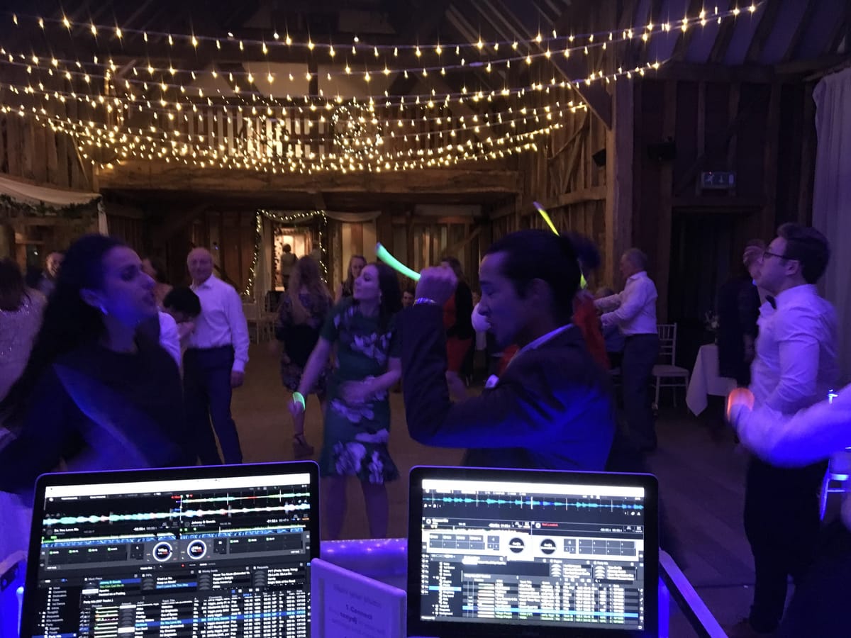View from behind the DJ decks of guests dancing under fairy lights late in a wedding evening
